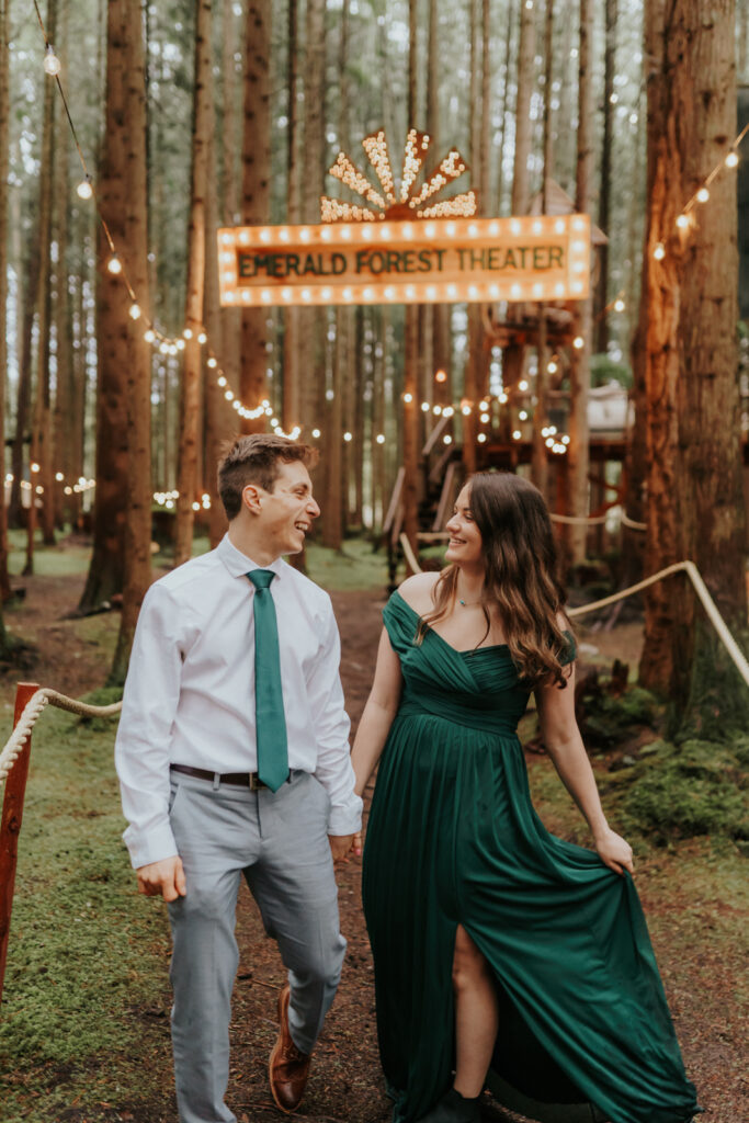 Photo of a couple walking in Emerald Forest Theater. The woman is wearing a beautiful green dress, and the man has a matching colored tie with a white shirt and grey pants that are well fitted.