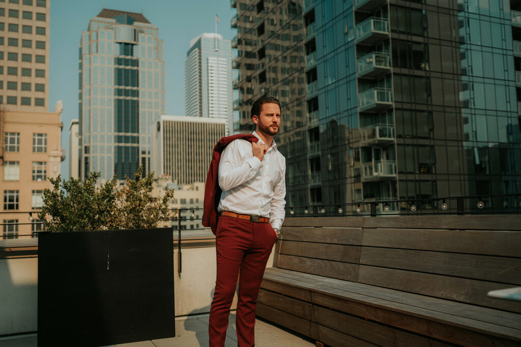 Groom in a red suit, white shirt at a rooftop in Seattle. Custom tailored suit with brown belt and shoes.