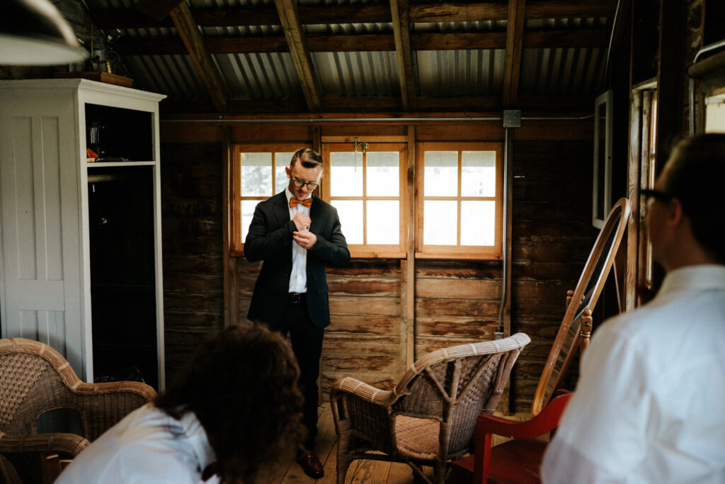 Photo of a groom getting ready in Seattle. He has a blue jacket on and is fixing his cuffs as his groomsmen watch him get ready