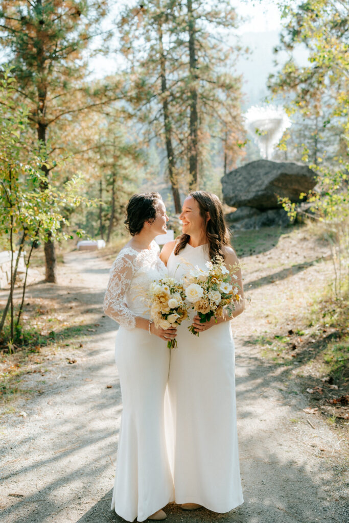 Photo of two Brides holding one another in Leavenworth, Washington as the look to each other both holding bouquets and smile to one another surrounded by trees and foliage