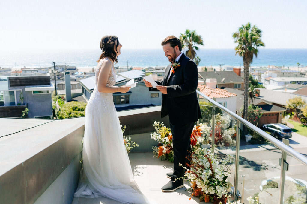 Photo of a Couple on a deck in Hermosa Beach, California. Bride and Groom reading their vows to one another with the ocean behind them while they laugh together.